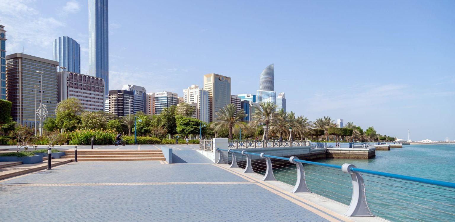 Abu Dhabi Corniche in the morning. People walking at the distance with modern skyscapers at the background. Abu Dhabi Corniche in the morning. High rise modern buildings at the background and seaside on the left. People walking and riding bikes at the distance. High quality photo.